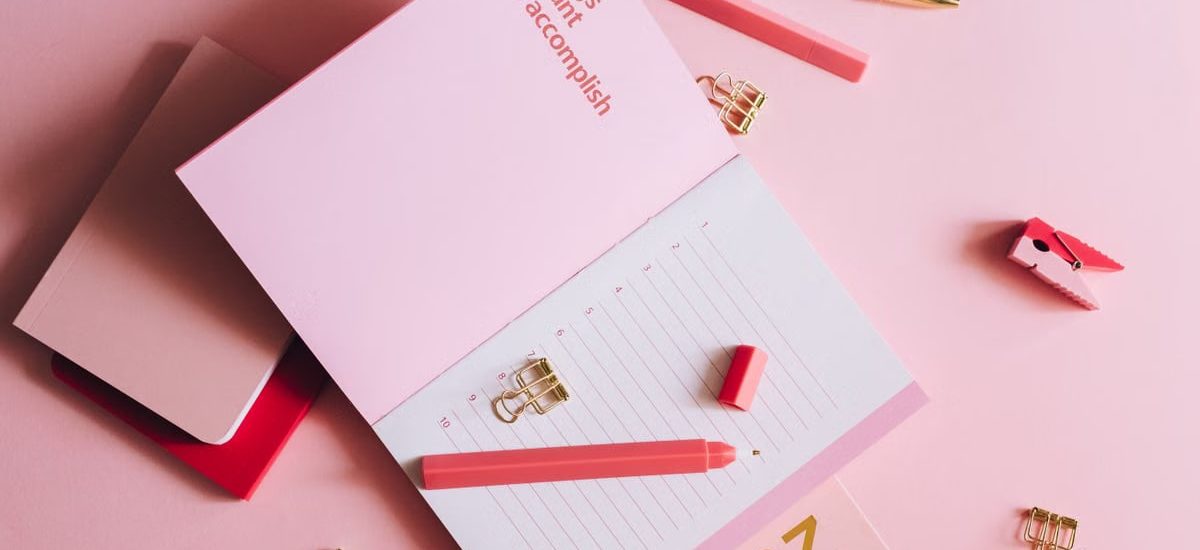 Organized workspace with pink planner, pen, and clips on pastel background.
